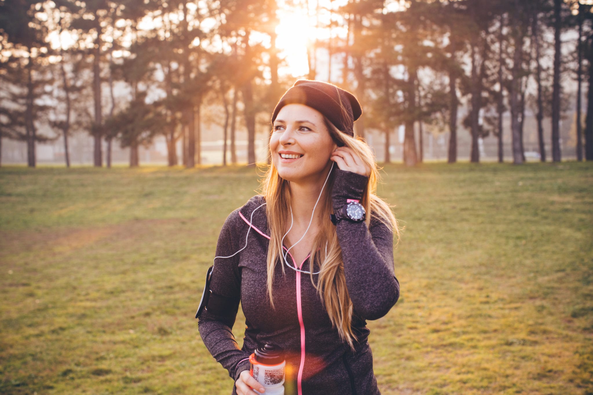 Young woman jogging outside in sunny autumn forest. She took a break to set up smartphone to monitor her progress or set up music and drink water. The sun is shining in the background .